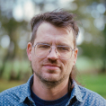 Portraitfoto eines Mannes mit Brille, hellen Haaren und blauem Hemd.