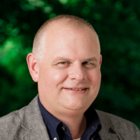 Matthias Emrich, Mann mit kurzen Haaren, ohne Bart, lächelt. Braunes Jacket und dunkles Hemd.