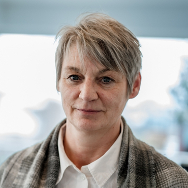 Portraitfoto von Andrea Burs, Leiterin Linientechnik bei LWLcom, Frau mit kurzem, grau-blondem Haar, weißem Hemd und kariertem Schal über den Schultern vor unscharfem Hintergrund.