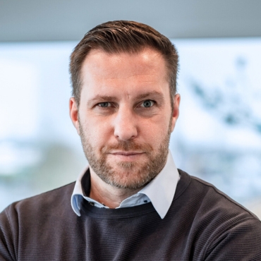 Portraitfoto von Mirko Dettmann, Leiter Vertrieb bei LWLcom, trägt ein hellblaues Hemd unter einem dunkelgrauen Pullover, sitzt vor einem Fenster mit unscharfem Hintergrund. 