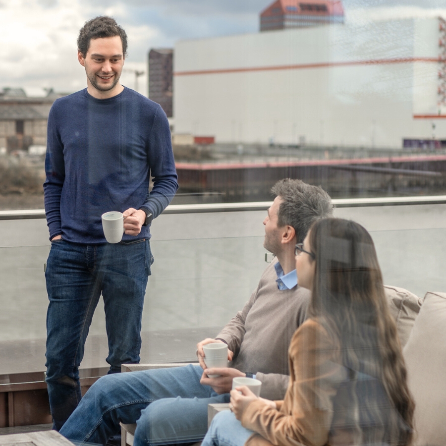 Ein Kollege und eine Kollegin sitzen mit einer Kaffeetasse auf der Dachterrasse und unterhalten sich mit einem weiteren Kollegen, der neben der Bank mit einer Kaffeetasse in der Hand steht.